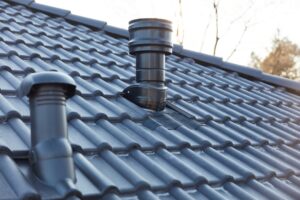 Close-up of a dark tiled roof with two black metal ventilation pipes extending upwards, photographed in natural daylight—ideal roof ventilation for homes exposed to coastal air.