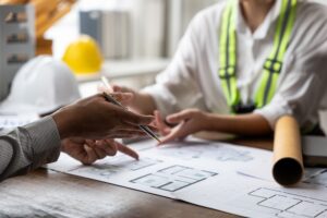 Two people discuss architectural blueprints at a desk, with construction helmets, a rolled-up plan, safety gear, and a Portland roofing permit checklist visible.
