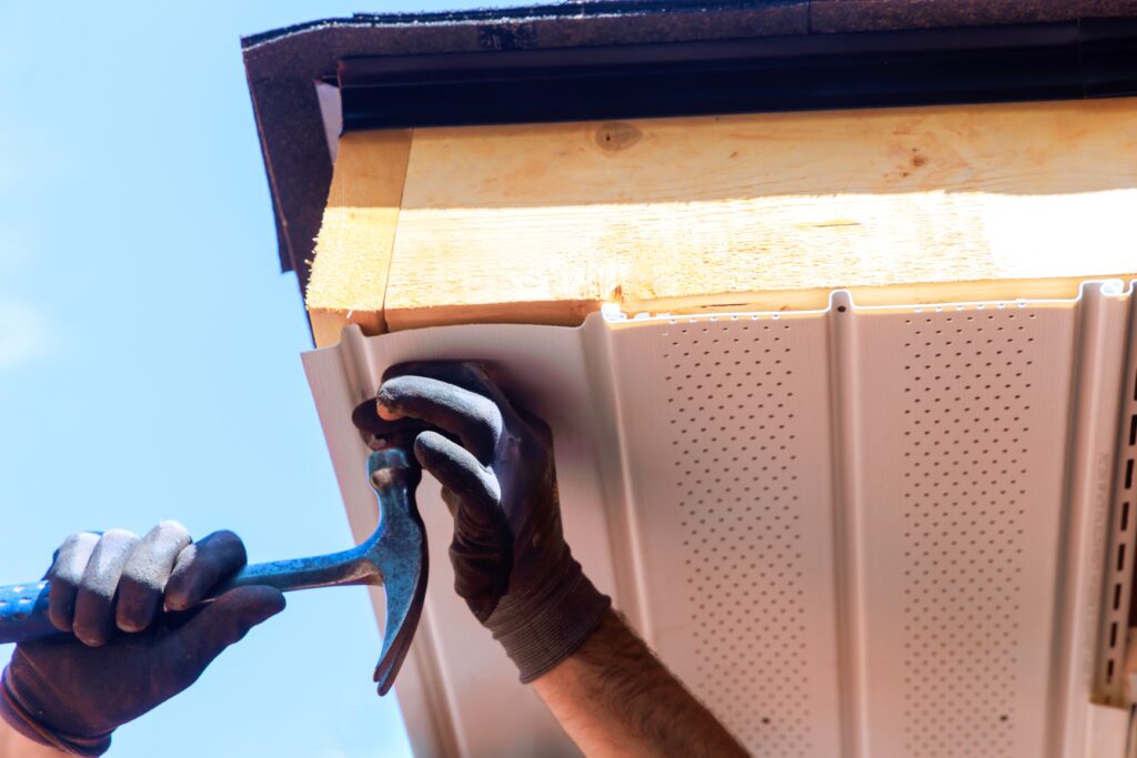 Person wearing gloves installs soffit panel under roof eave, using a hammer to secure it, while checking for signs of roof flashing failure, with blue sky in the background.