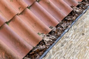 A roof with red clay tiles and a gutter filled with dry, fallen leaves runs along the edge of a stone wall, highlighting the need for regular gutter maintenance to prevent water damage in Portland homes.