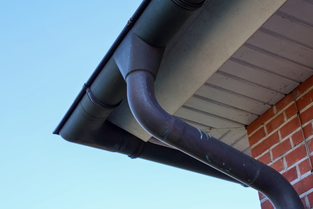 Close-up view of a brown metal gutter and downspout attached to the edge of a roof on a brick building against a clear blue sky, highlighting the importance of gutter maintenance for preventing water damage to Portland homes.