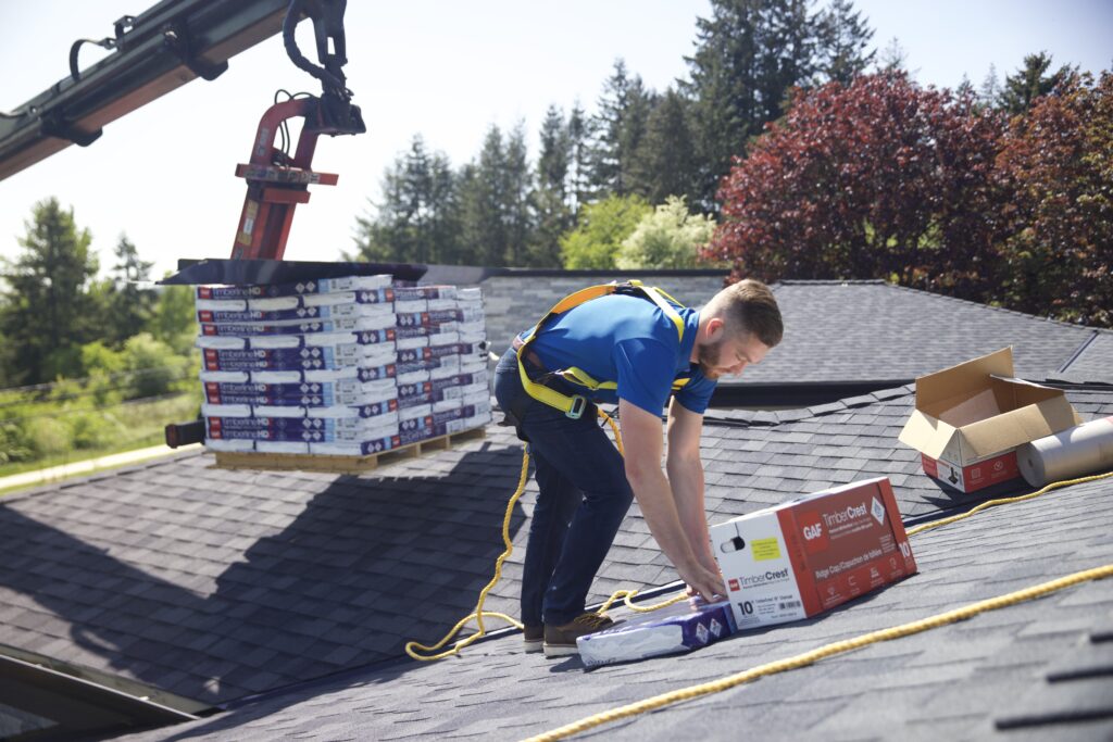A man wearing safety gear works on a roof, handling roofing materials while a crane delivers more packages nearby. As part of the Blue Moon Roofing team, he ensures every detail is managed expertly, from shingles to gutters. Trees and other rooftops are visible in the background.