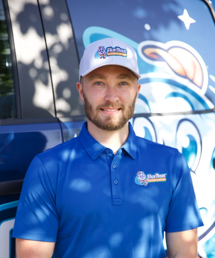 Man wearing a Blue Moon uniform stands in front of a branded vehicle with the Blue Moon logo visible on his shirt, hat, and the van. Grayson Carey, President of Blue Moon Roofing, exemplifies professionalism and company pride in this image.