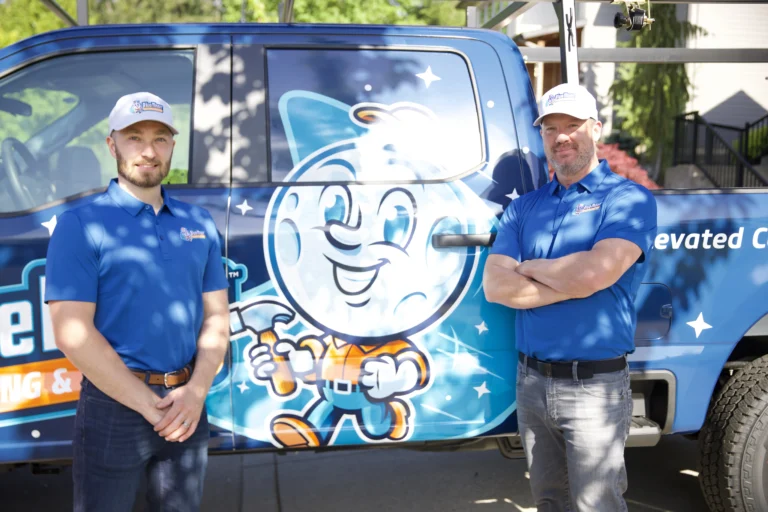 Two men in blue polo shirts and hats stand next to a blue work truck featuring a cartoon mascot and company branding on its side.
