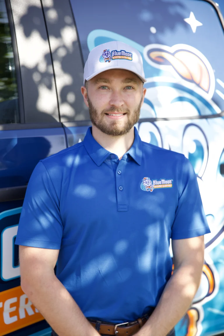 A man in a blue “Blue Moon” company shirt and cap stands in front of a branded vehicle, smiling at the camera.