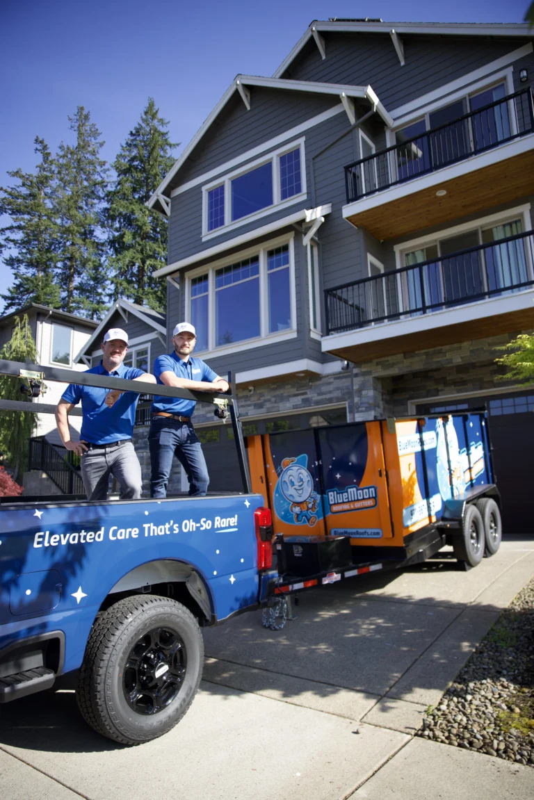 Two workers in blue uniforms stand on a Blue Moon service truck parked outside a modern multi-story gray house with balconies.