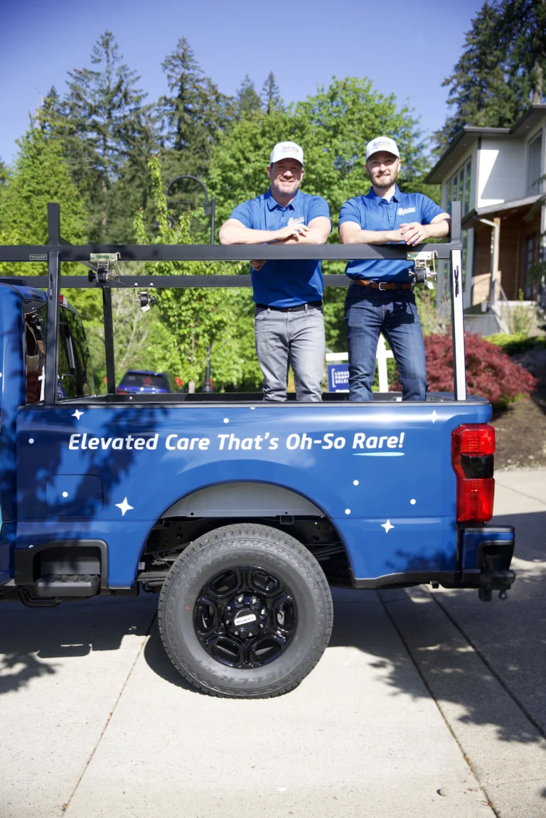 Two men in blue shirts and hats stand in the back of a blue truck with the slogan “Elevated Care That’s Oh-So Rare!” parked in a residential driveway on a sunny day.