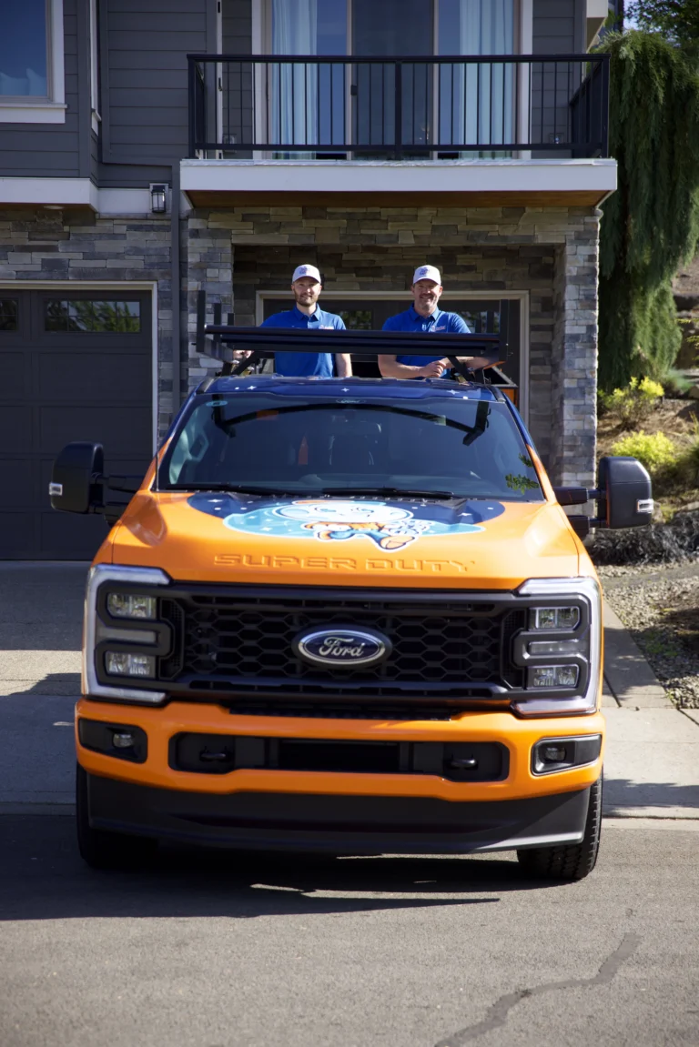 Two people in blue shirts and hats stand in the bed of an orange Ford truck with a cartoon graphic, parked in front of a house.