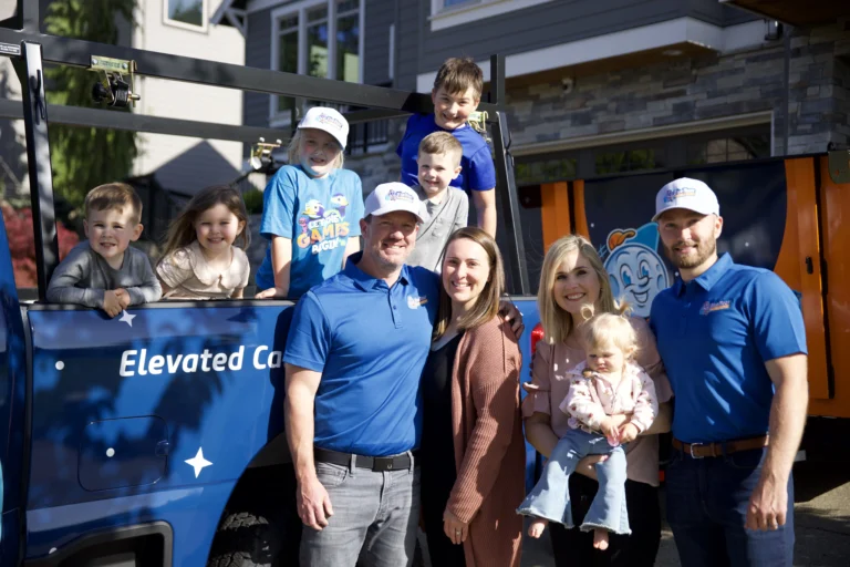 A group of adults and children pose and smile in front of two blue trucks with “Elevated” signage in a residential driveway on a sunny day.