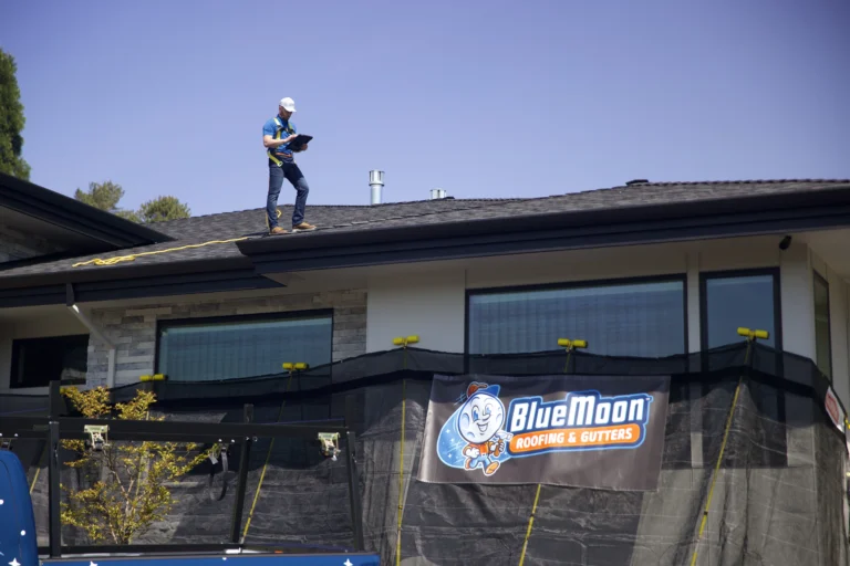 A worker stands on a house roof holding a clipboard. Below, a sign reads “Blue Moon Roofing & Gutters.” The house is under renovation with safety barriers in front.