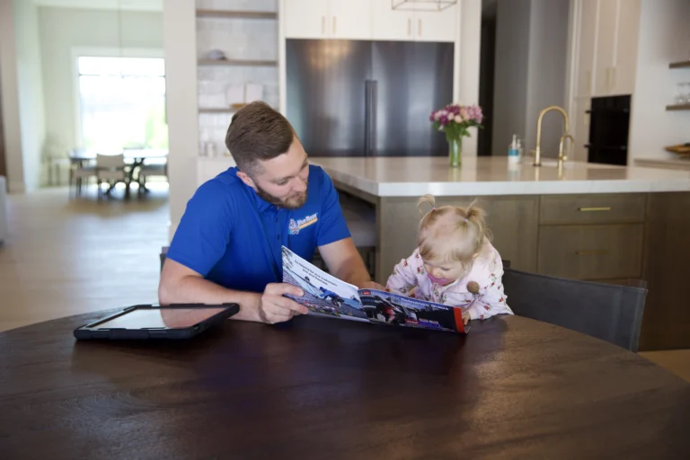 A man in a blue shirt reads a picture book with a young child at a round table in a modern kitchen.