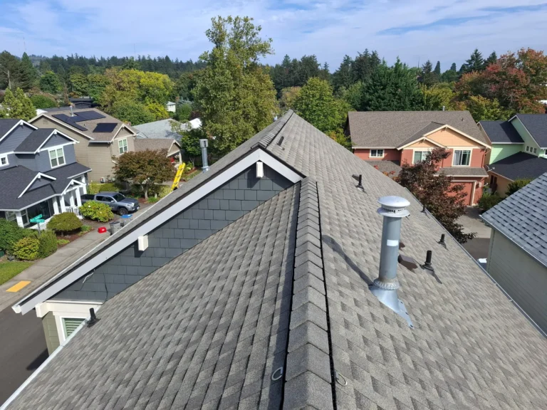 View from the peak of a residential roof, showing gray shingles, vent pipes, and a suburban neighborhood with trees and houses in the background.