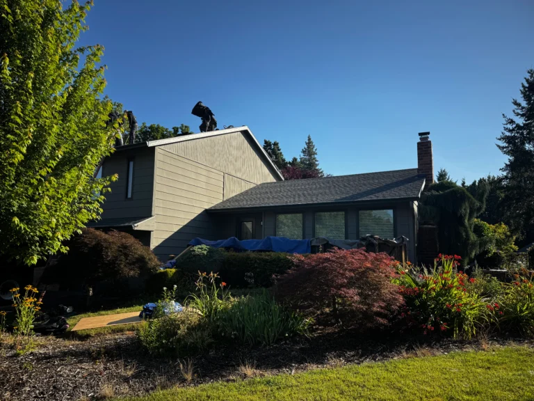 A person stands on the roof of a two-story house surrounded by trees and plants under a clear blue sky.