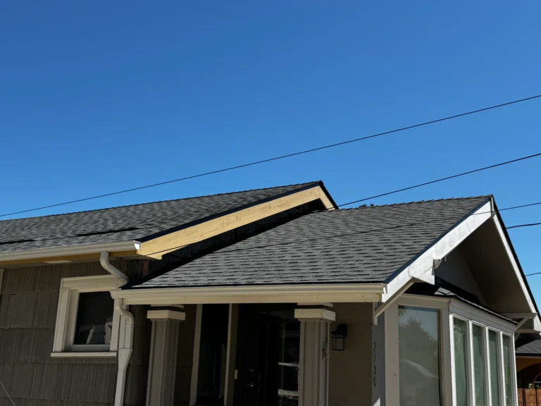 A house with dark gray shingles and exposed wood trim on the roof under a clear blue sky, with visible gutters and power lines.