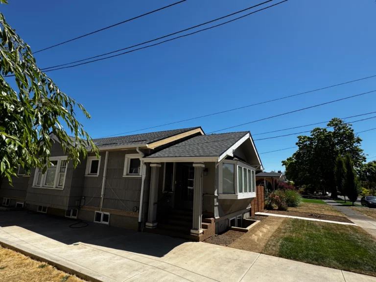 Single-story house with gray siding, a covered porch, and a manicured lawn on a sunny day, located at the corner of a residential street.